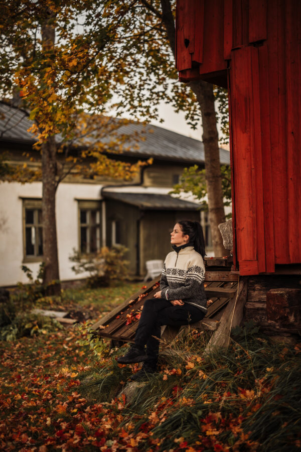Syksyinen otos naisesta, joka istuu rentoutuneena vanhan maalaistalon pihalla, yllään kuvioitu villapaita. Taustalla näkyy keltaisten ja oranssien lehtien koristama puu, mikä luo lämpimän tunnelman viileään päivään.